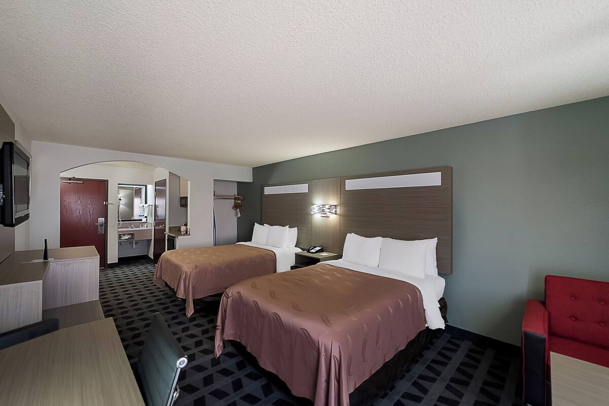A hotel room with two beds, a desk, a chair, a TV, and a bathroom visible through an open door.