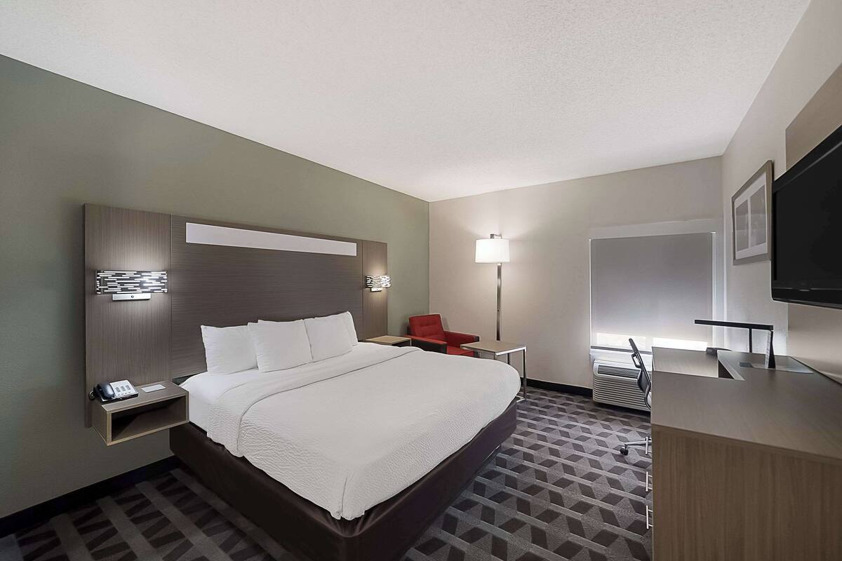A hotel room with a large bed, bedside table with a phone, a red chair, a desk with a lamp, and a TV on the wall.