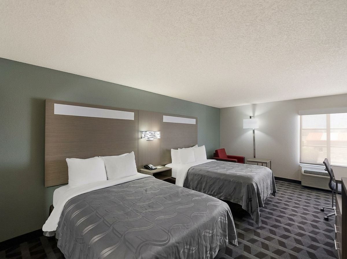 A hotel room with two beds, a desk, a chair, a lamp, and a window.