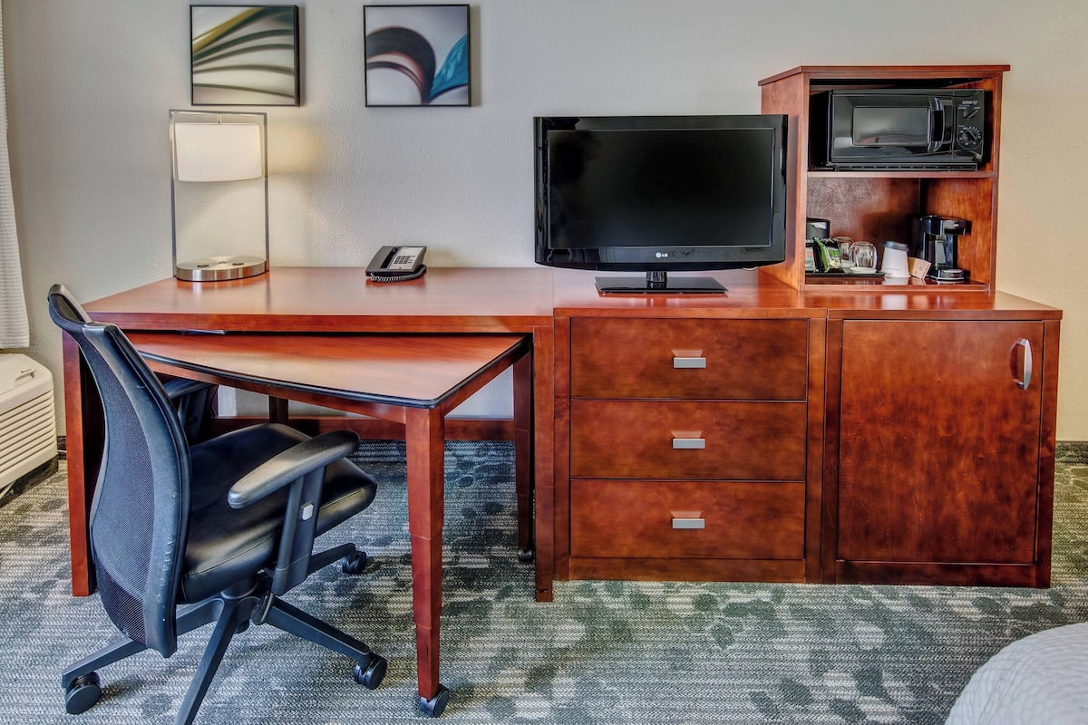 A hotel room with a wooden desk, a black office chair, a flat-screen TV, a microwave, a coffee maker, and a telephone.