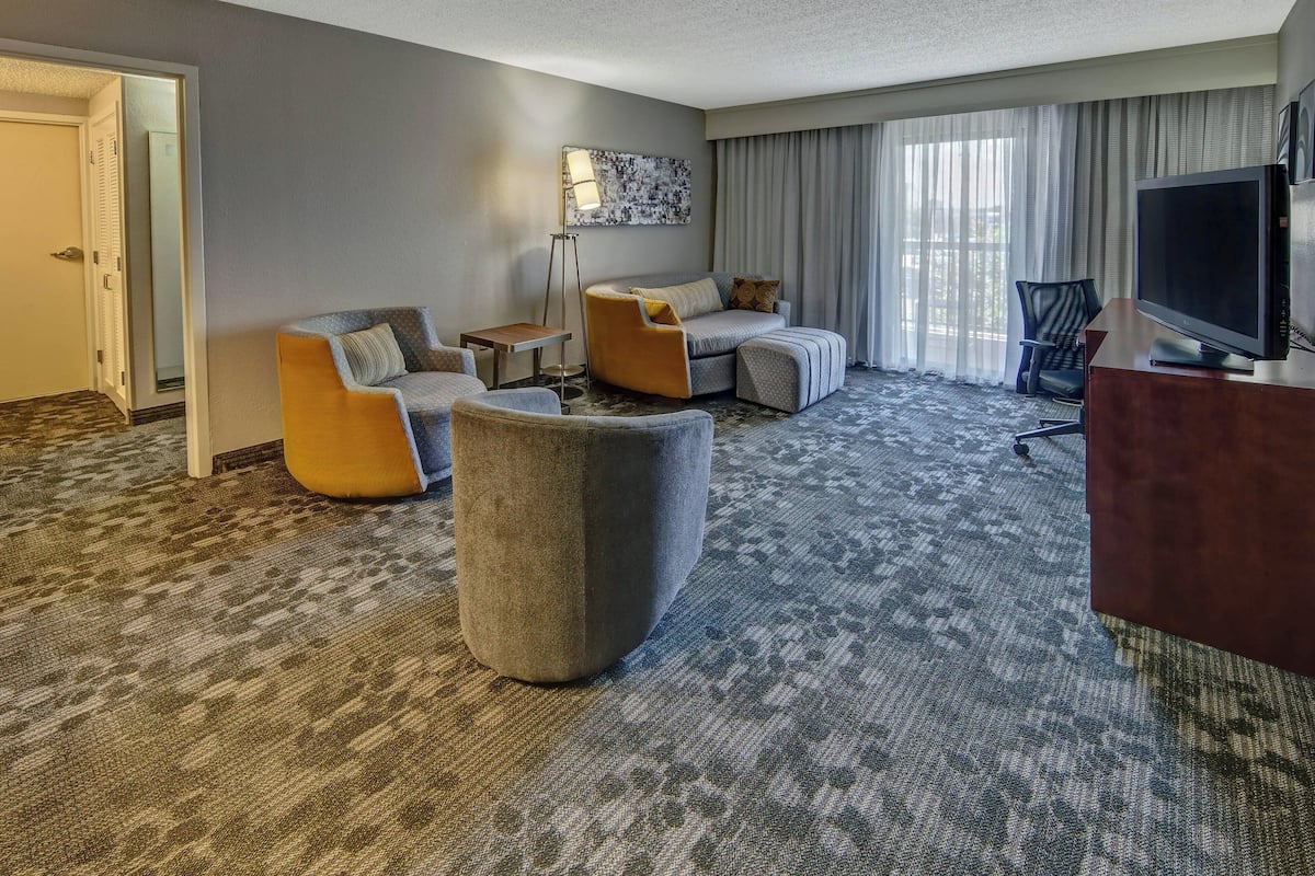 A hotel room with a grey carpet, a sofa set, a TV, and a large window with curtains.