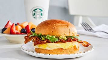 Un sándwich de desayuno con tocino, lechuga y huevo en un pan, un plato de fruta y una taza de Starbucks al fondo.