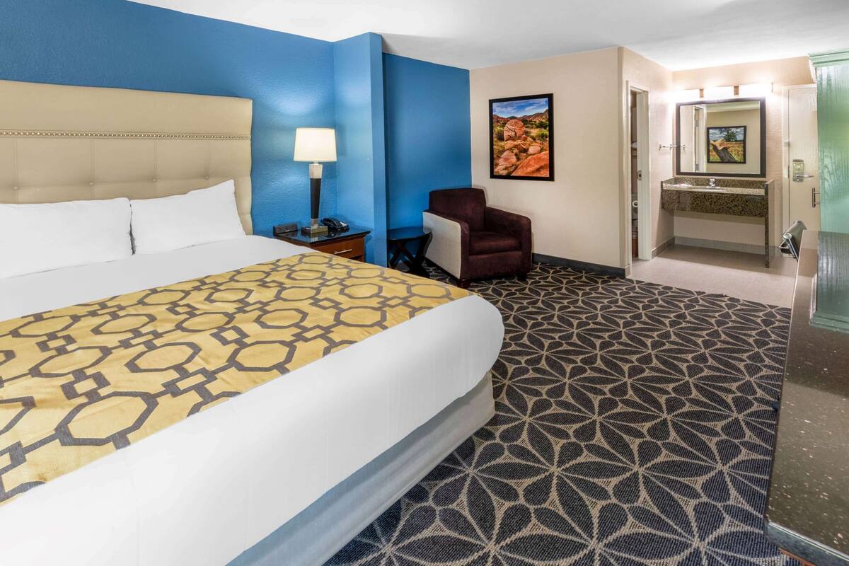 Habitación de hotel con una cama grande, un escritorio con lámpara, una silla, un baño con lavamanos y espejo, y una alfombra estampada.