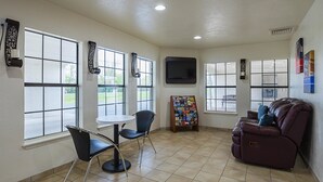 Una habitación amplia con ventanas grandes, un televisor de pantalla plana en la pared, una pequeña mesa redonda, dos sillas negras, un sofá de cuero marrón con cojines azules y un tablón de anuncios con varios objetos.