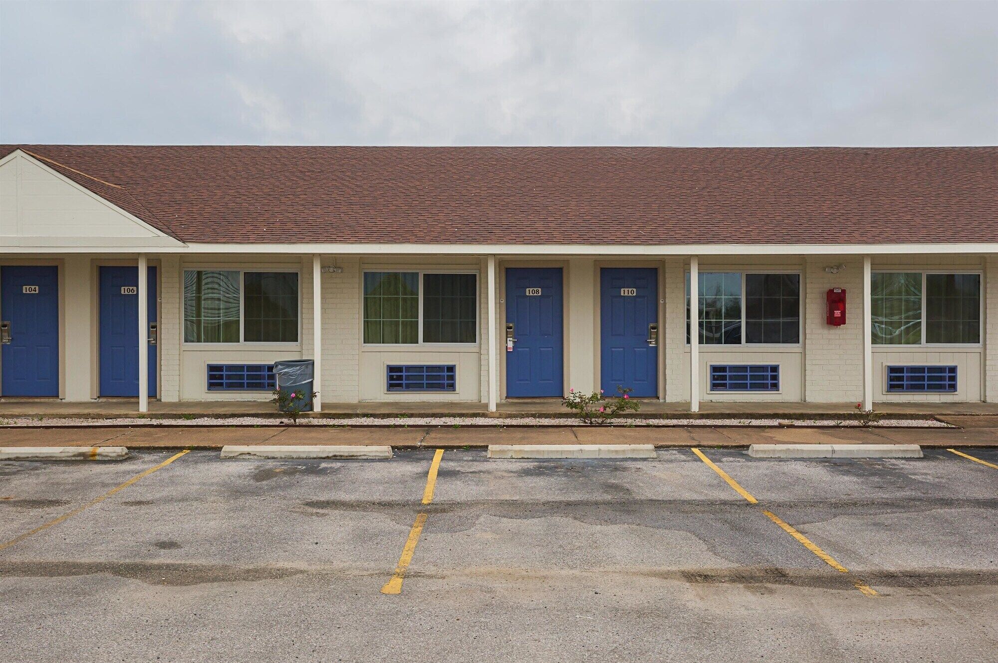 Un motel con seis puertas azules numeradas del 104 al 110, un buzón rojo y un estacionamiento con líneas amarillas.