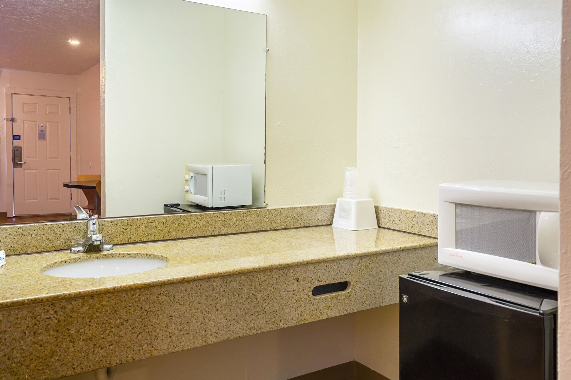 Un baño con lavamanos de encimera de granito, espejo, microondas y un refrigerador pequeño.