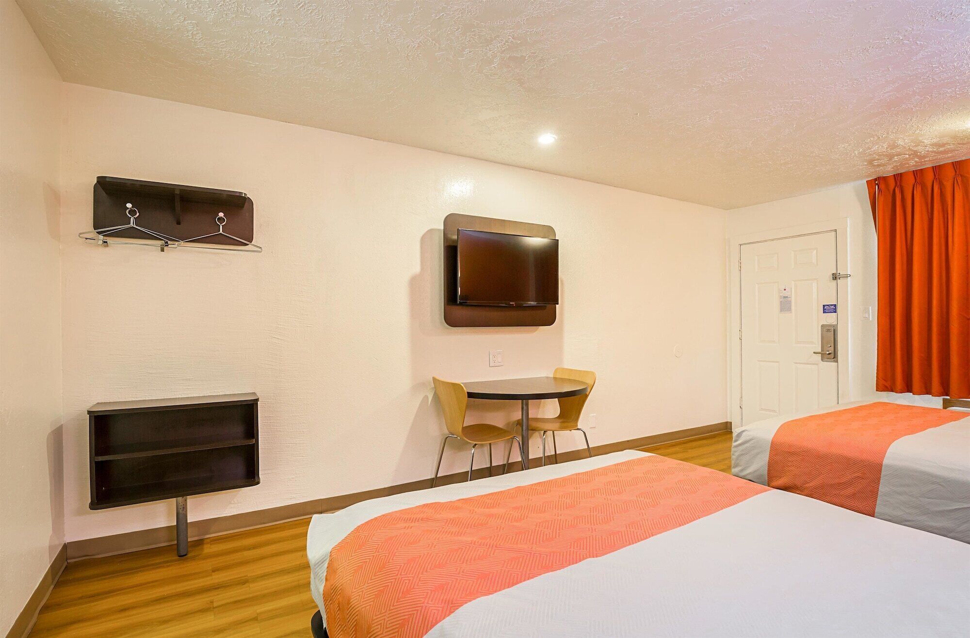 A hotel room with two beds, a TV mounted on the wall, a small table with chairs, and a coat rack.