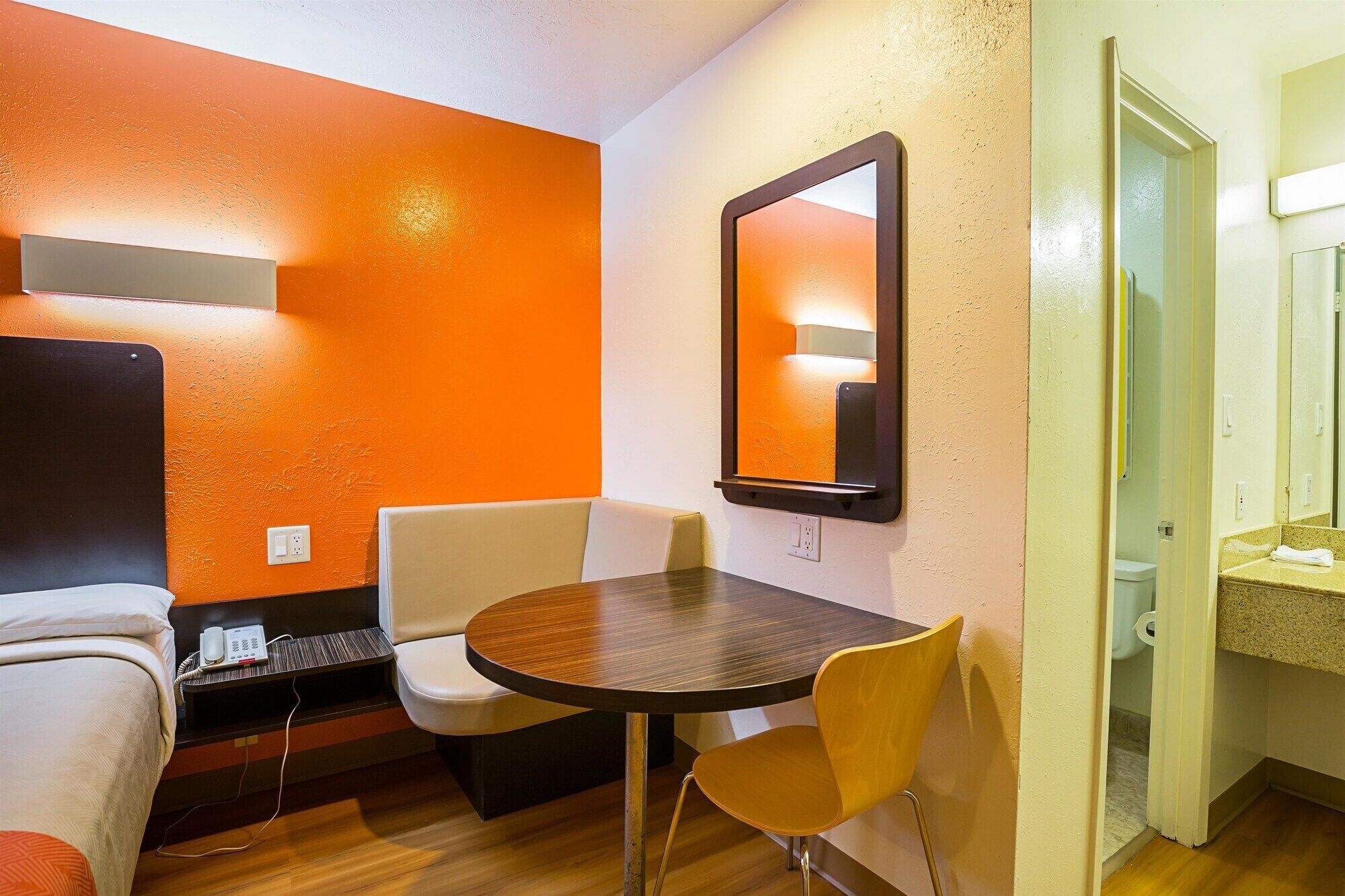 A hotel room with an orange accent wall, a round table, two chairs, a bed, a small desk with a phone, and a mirror.