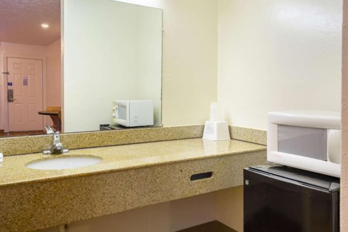 Un baño de hotel con lavamanos de encimera de granito, espejo, inodoro blanco y microondas.