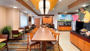 A hotel dining area with a long wooden table, multiple chairs, a kitchen area with a refrigerator, and a mural on the wall. - Fairfield Inn & Suites by Marriott® Dallas Plano West (Plano)