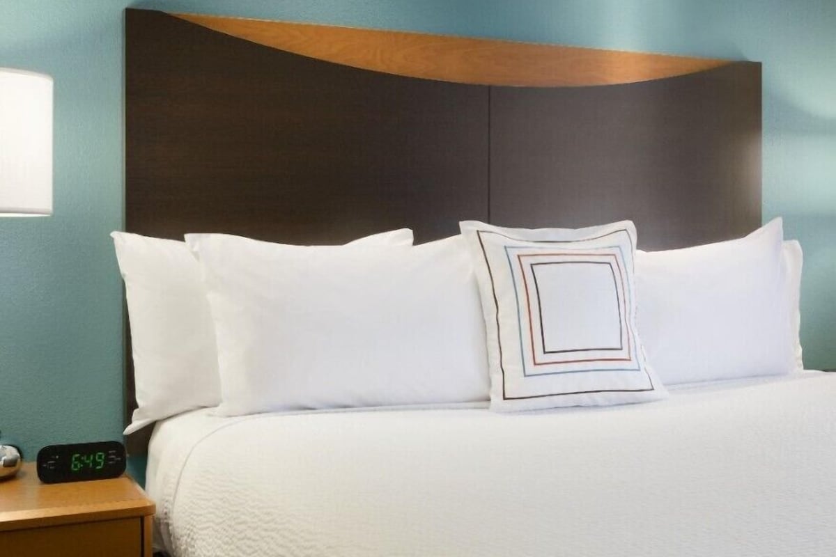 A neatly made bed with white linens and a geometric-patterned pillow, a wooden headboard, and a digital clock displaying the time.