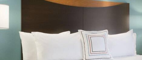 Una cama bien hecha con sábanas blancas y un cojín estampado con motivos geométricos, un cabecero de madera y un reloj digital que muestra la hora.