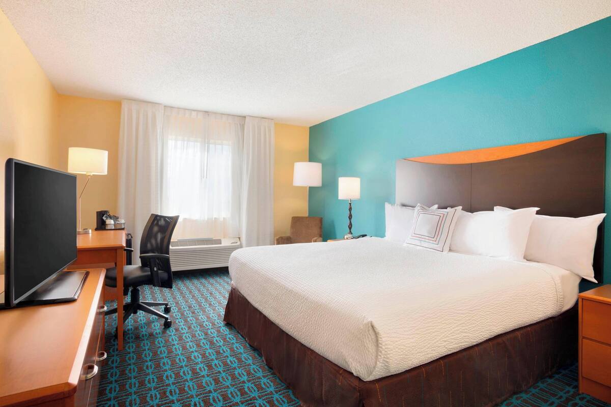A hotel room with a large bed, a desk with a TV, a chair, and a lamp.