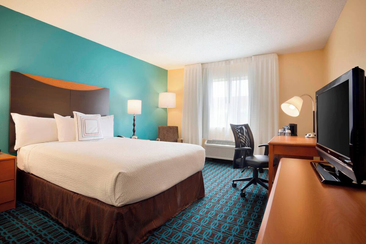 A hotel room with a large bed, a desk with a television, a chair, a desk lamp, and a window with curtains.