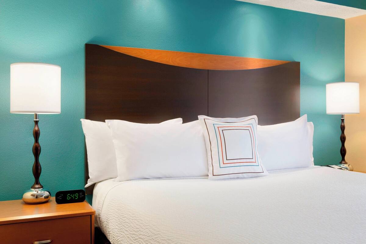 A hotel room with a bed, two bedside lamps, a digital clock, and teal walls.