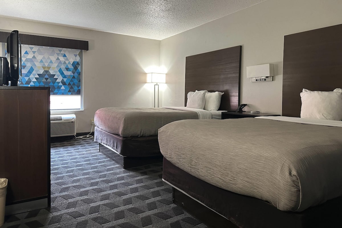 A hotel room with two beds, a desk, a lamp, and a wall-mounted air conditioner.