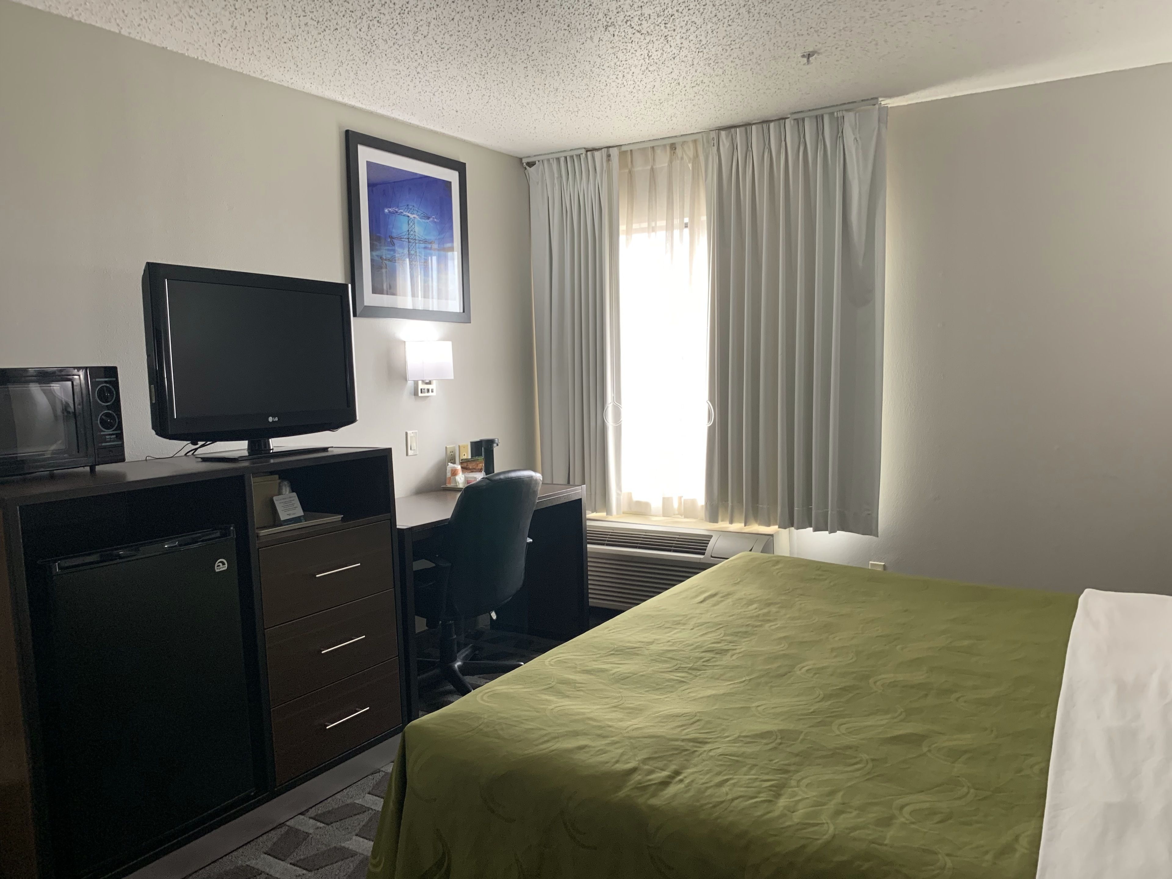 Une chambre d’hôtel comprenant un lit, un bureau avec une chaise, une télévision, un four à micro-ondes et une fenêtre avec des rideaux.