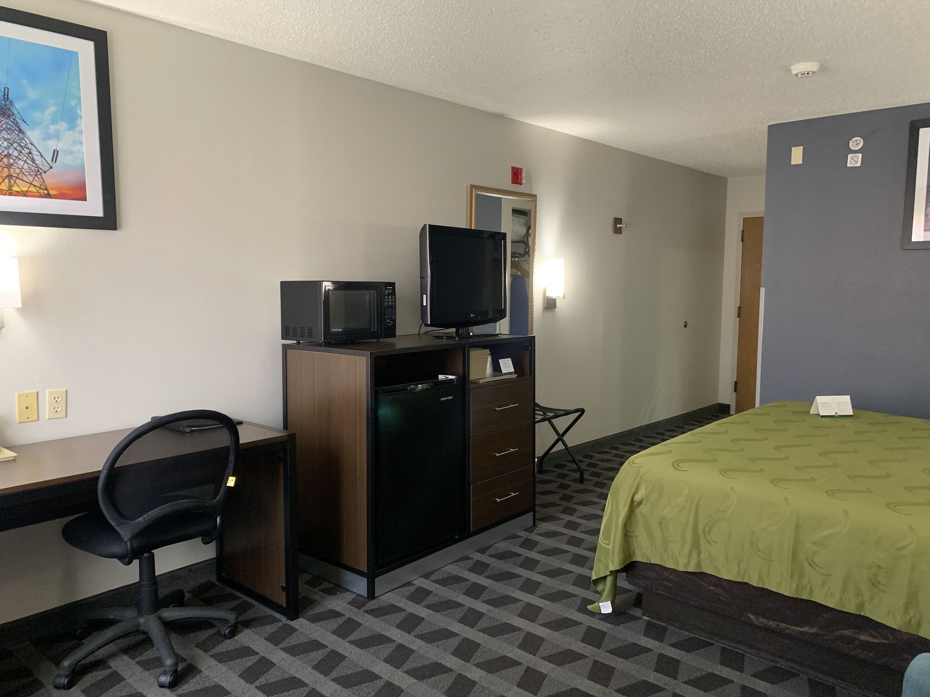 A hotel room with a desk, a chair, a bed with a green bedspread, a television on a dresser, and a painting on the wall.