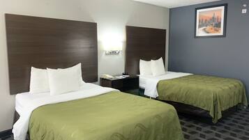 Habitación de hotel con dos camas, cada una con ropa blanca y almohadones verdes. Entre las camas, una mesita de noche con un teléfono y una lámpara. En la pared, un cuadro enmarcado de un paisaje urbano.