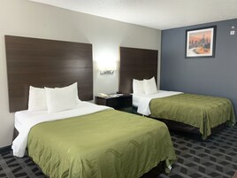 Habitación de hotel con dos camas, cada una con ropa blanca y almohadones verdes. Entre las camas, una mesita de noche con un teléfono y una lámpara. En la pared, un cuadro enmarcado de un paisaje urbano.