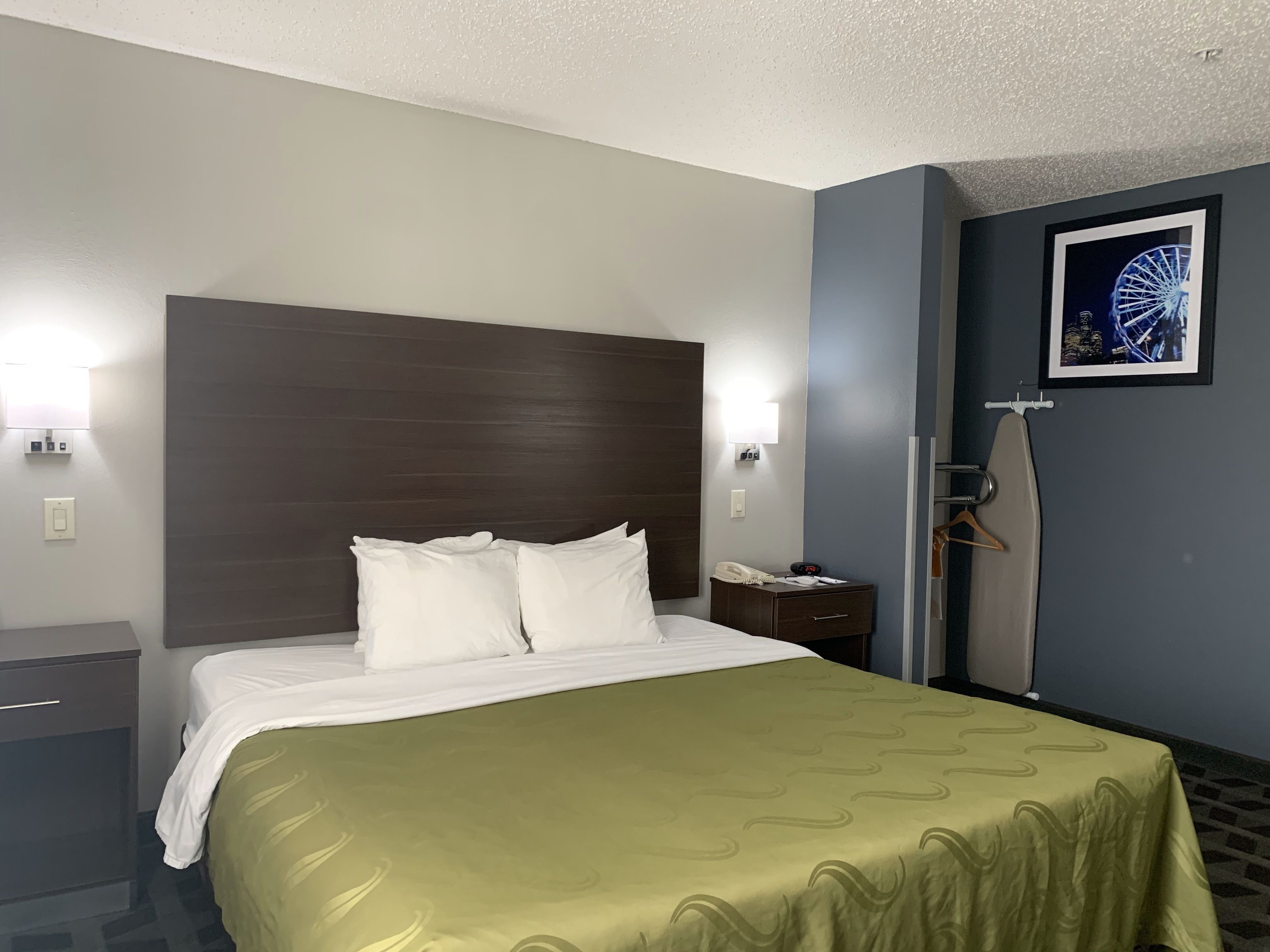 Habitación de hotel con una cama grande, cabecera de madera, dos lámparas de noche, una mesita de noche con un teléfono y una lámpara, un perchero y un cuadro en la pared.