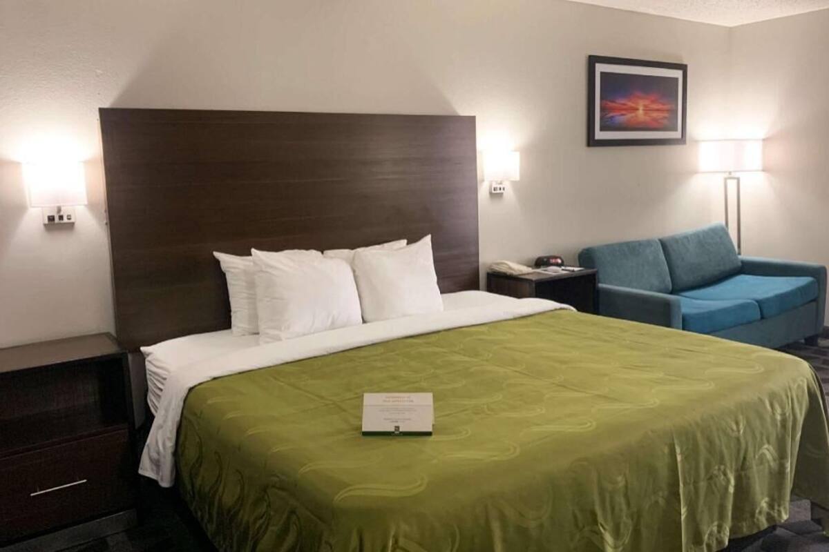 A hotel room with a bed, a green bedspread, a blue armchair, a nightstand, a lamp, and a framed picture on the wall.