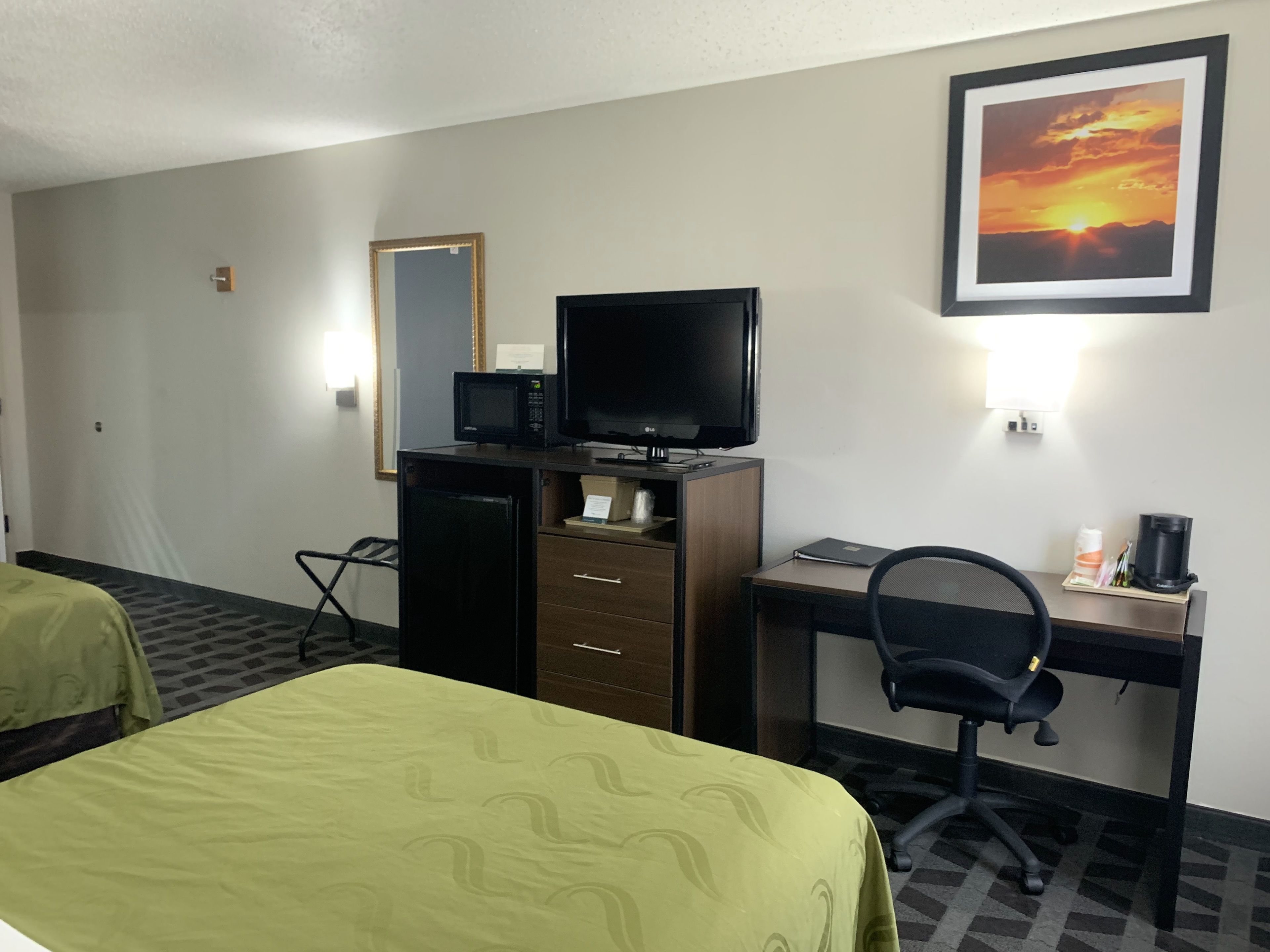 A hotel room with a bed, a desk with a computer and a TV, a small fridge, a chair, and a framed picture on the wall.