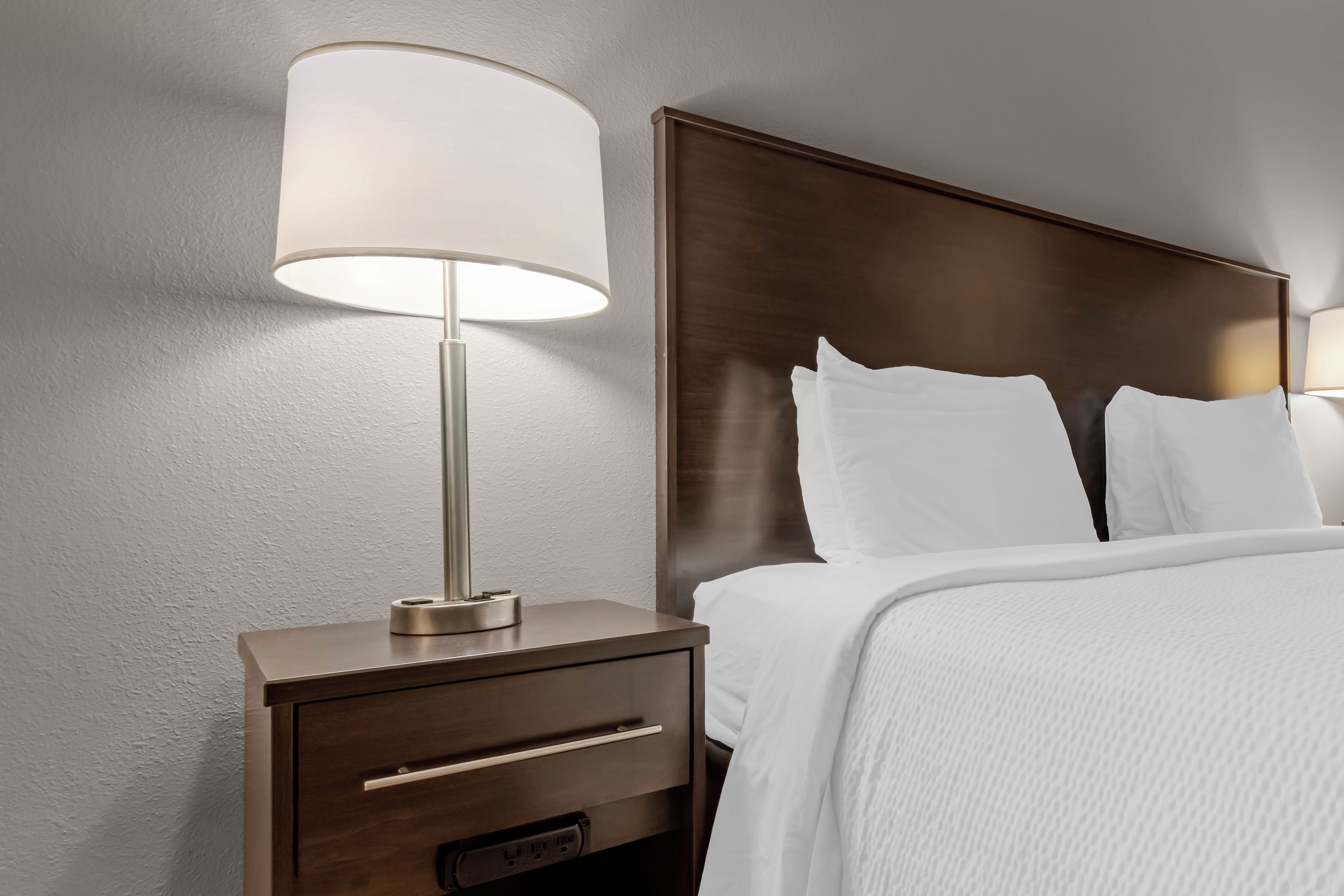 Quarto de hotel com cama, mesa de cabeceira com abajur e gaveta.