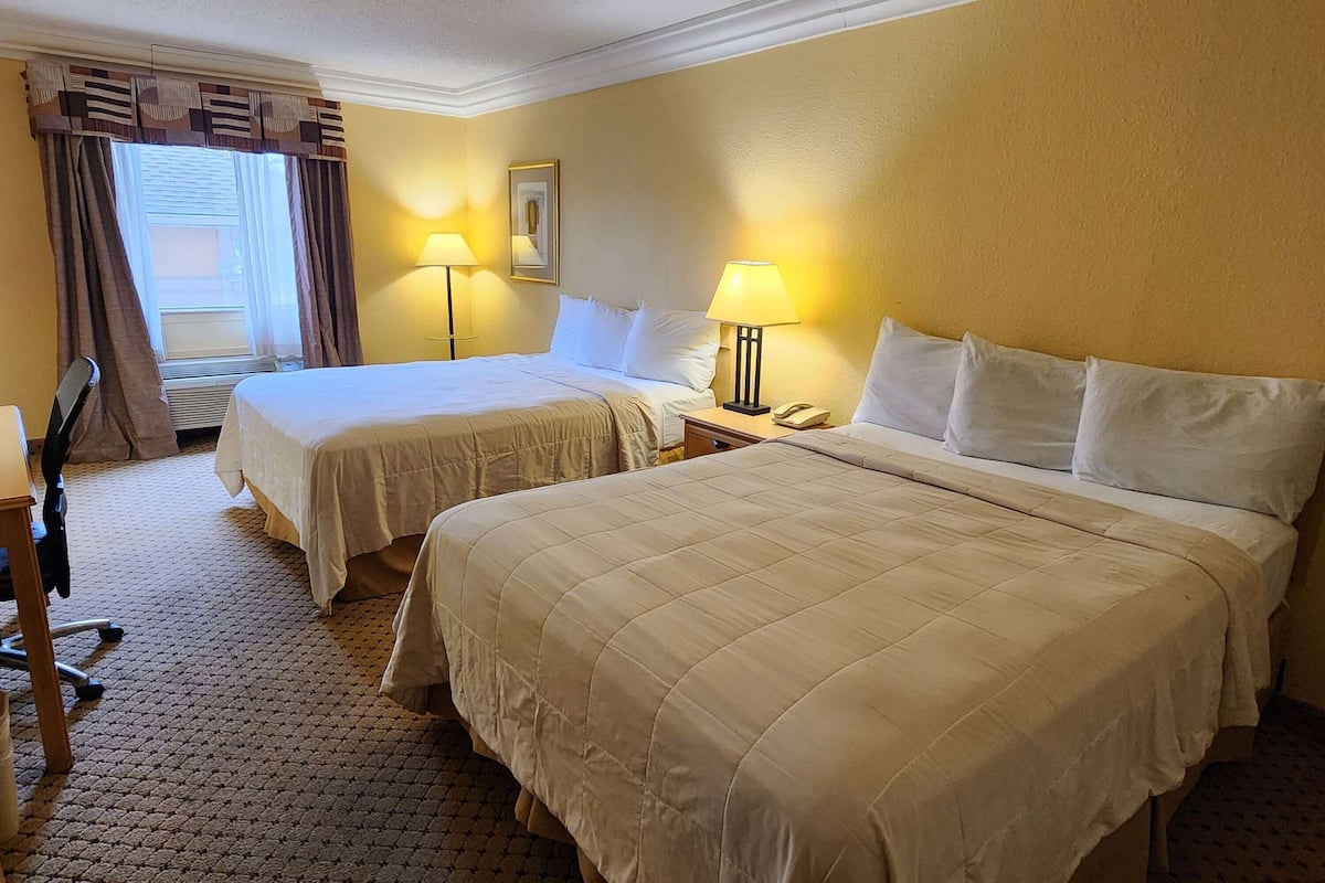 A hotel room with two beds, a desk, and a chair. There are two bedside lamps and a window with curtains.