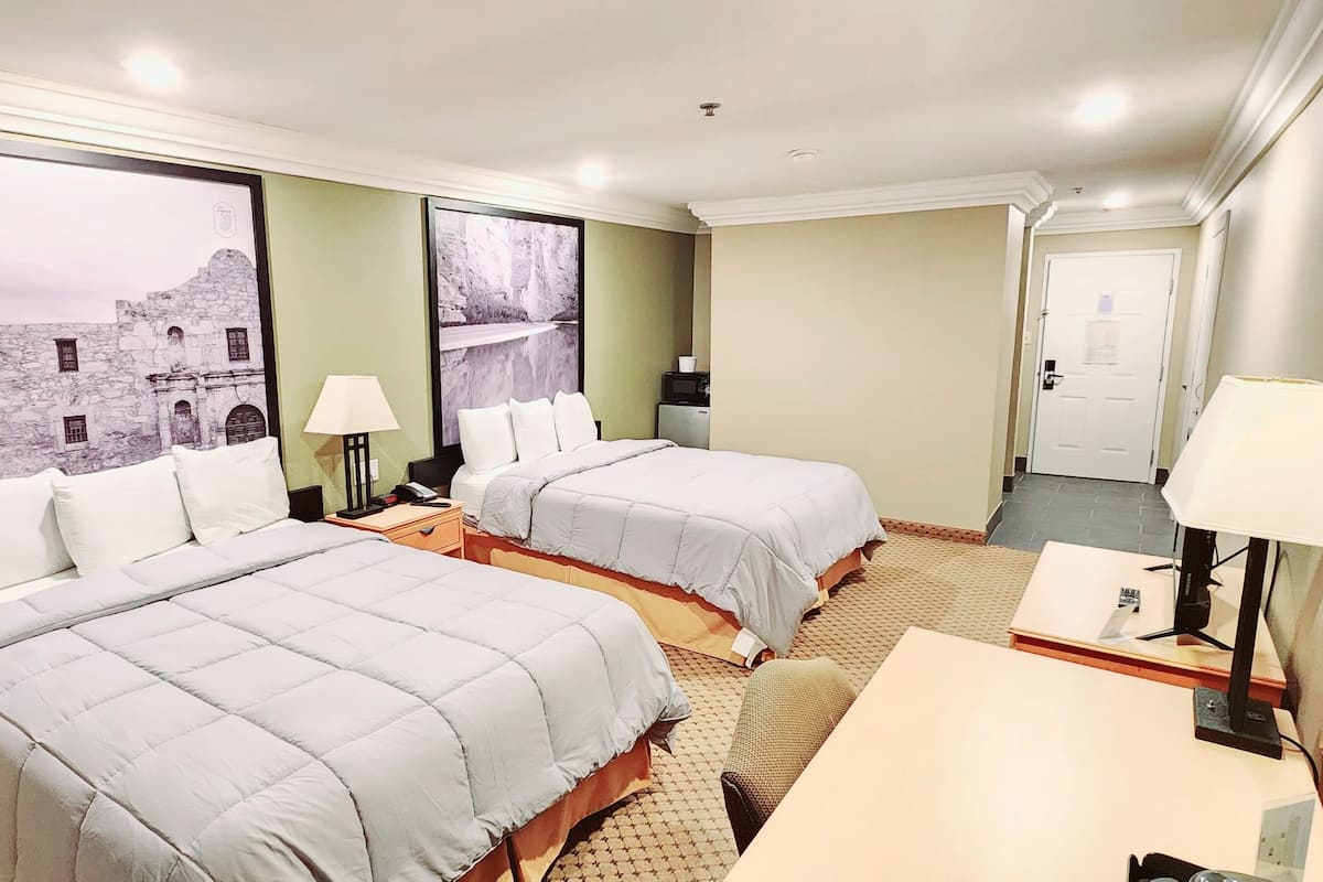 A hotel room with two beds, a desk, and a chair. There are two lamps on the desk and a small table. The walls have large framed pictures.