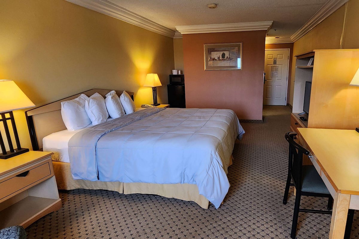 A hotel room with a large bed, bedside tables with lamps, a desk with a chair, a television, and a door leading to another room.