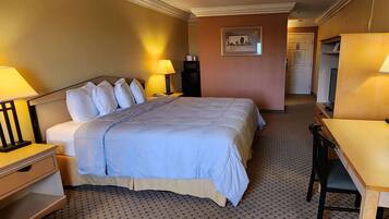 A hotel room with a large bed, bedside tables with lamps, a desk with a chair, a television, and a door leading to another room.