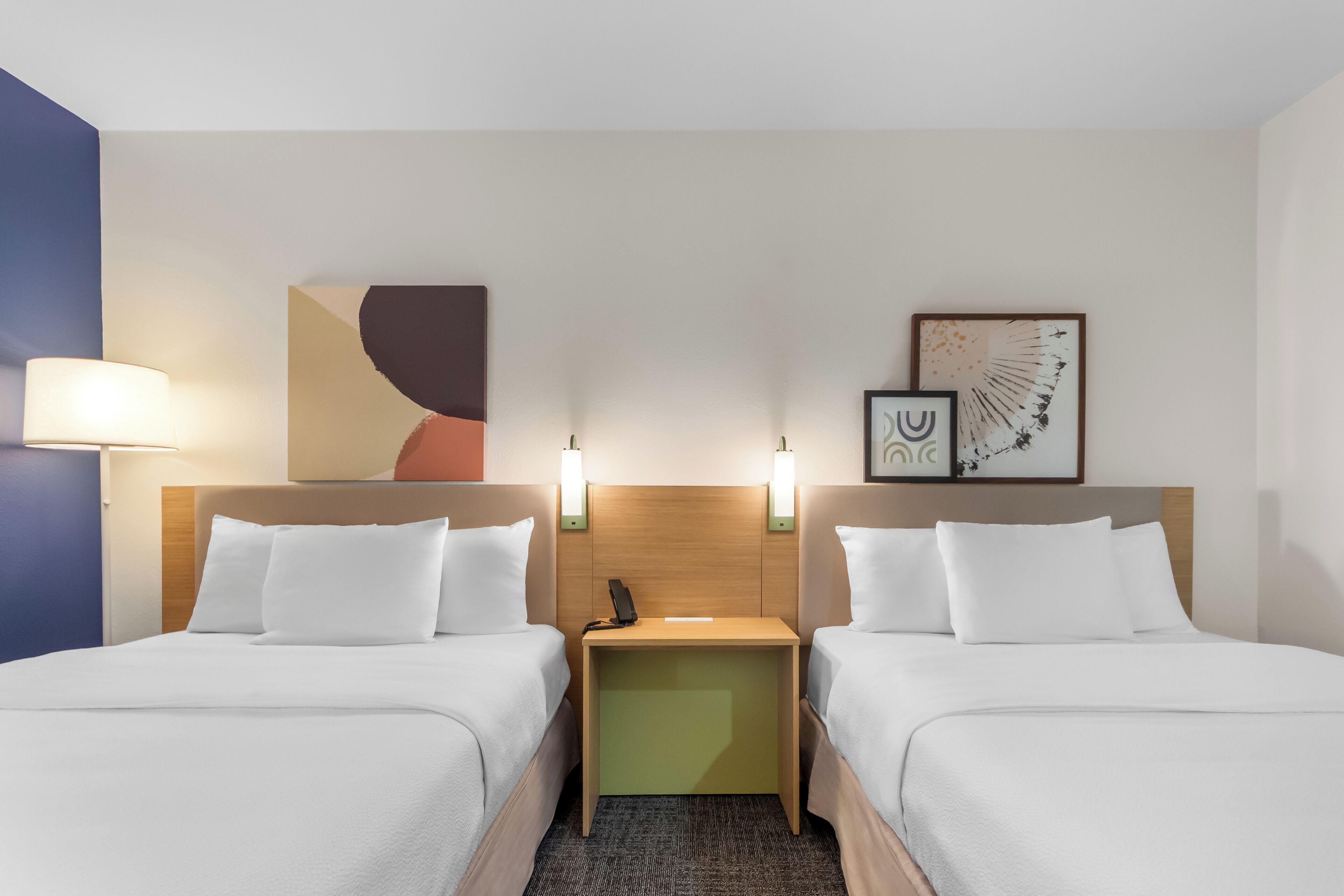 A hotel room with two beds, a nightstand, wall art, and a lamp.