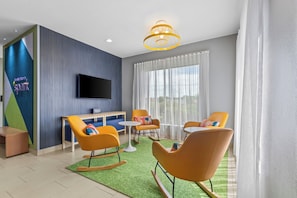 A modern hotel room with a flat-screen TV, orange rocking chairs, a small table, and a green rug.