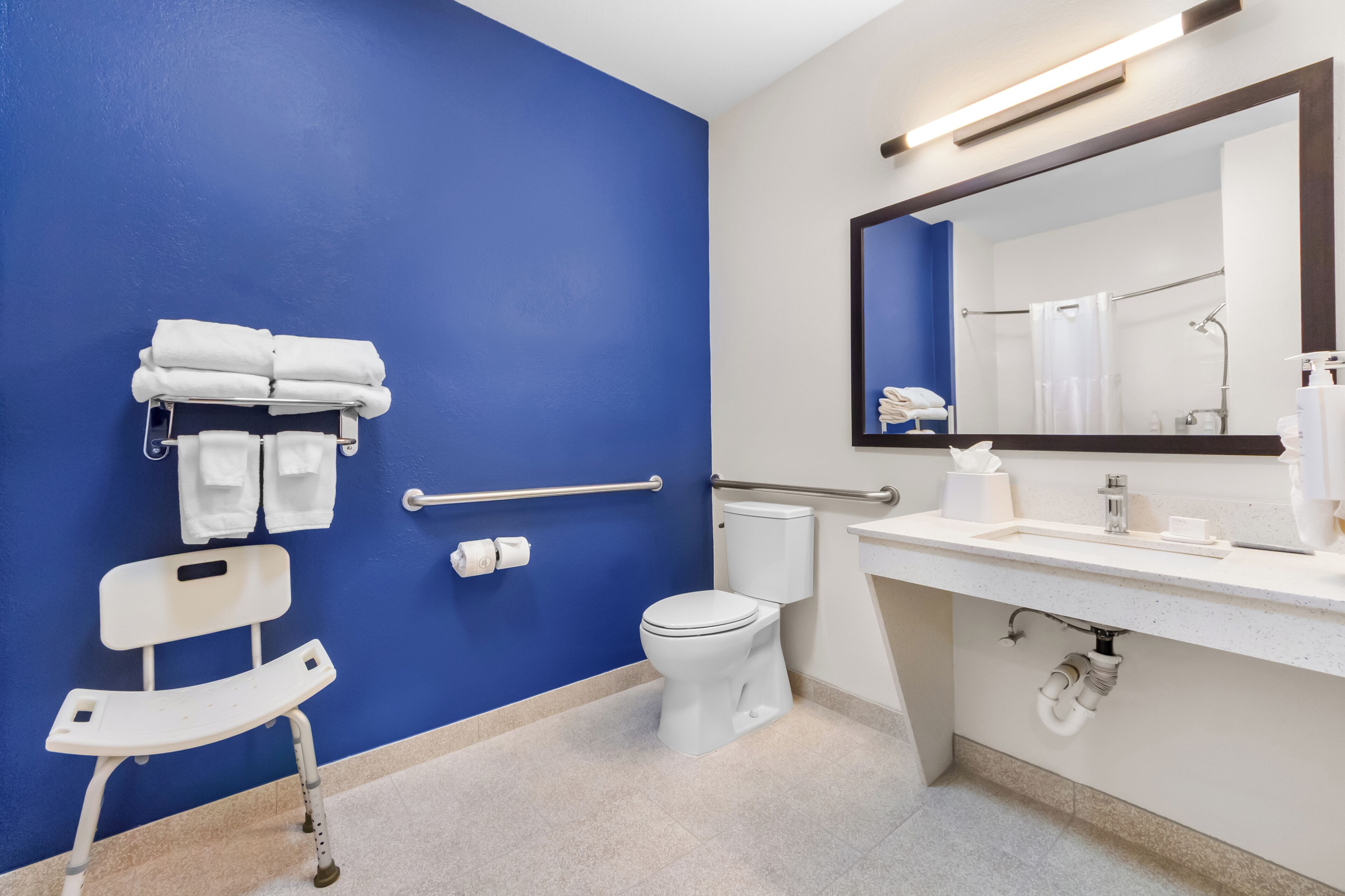 Un baño con pared azul, sanitarios blancos, un banco accesible para sillas de ruedas, un inodoro y un lavamanos con espejo.