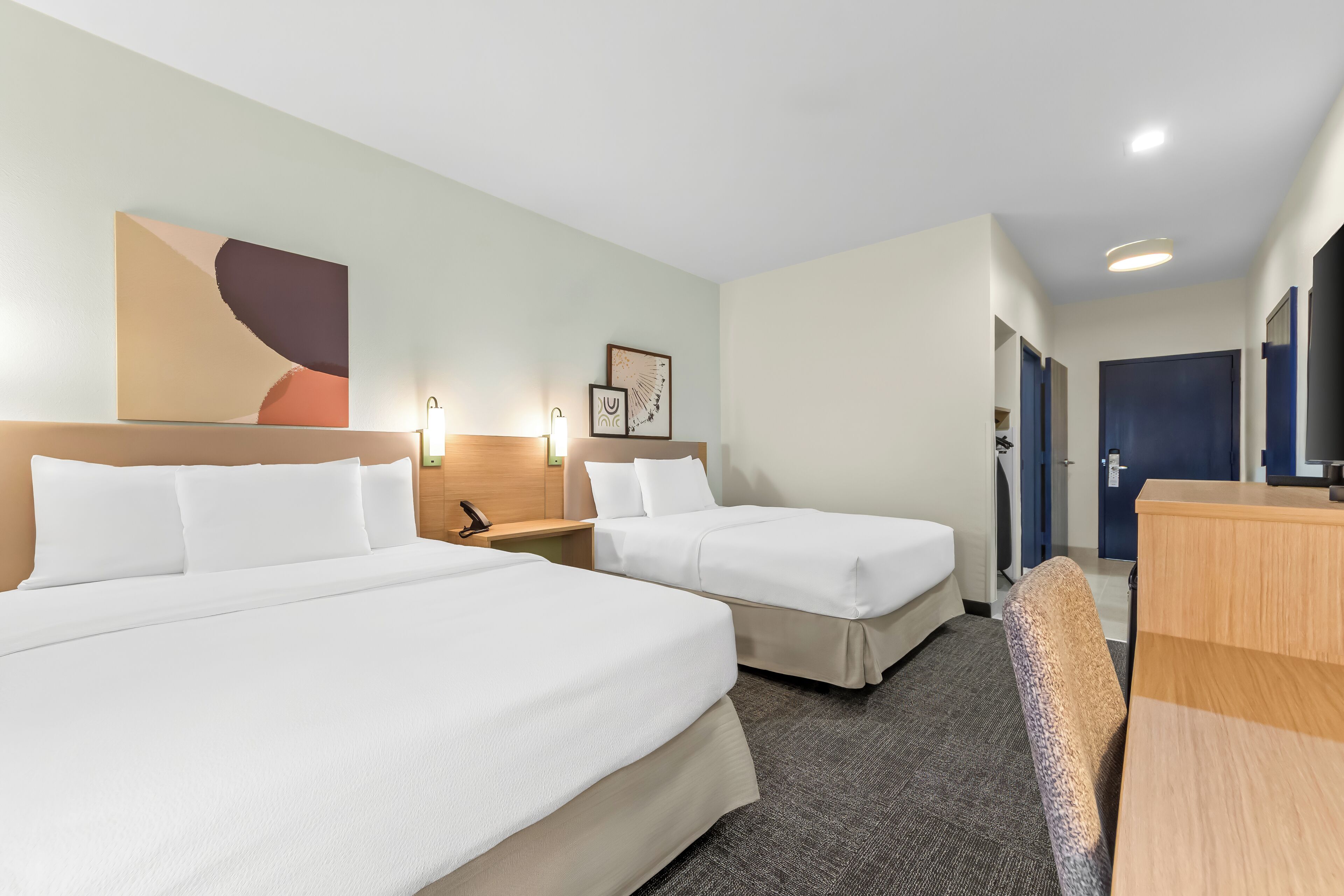 A hotel room with two beds, a desk, a chair, a lamp, and a painting on the wall.