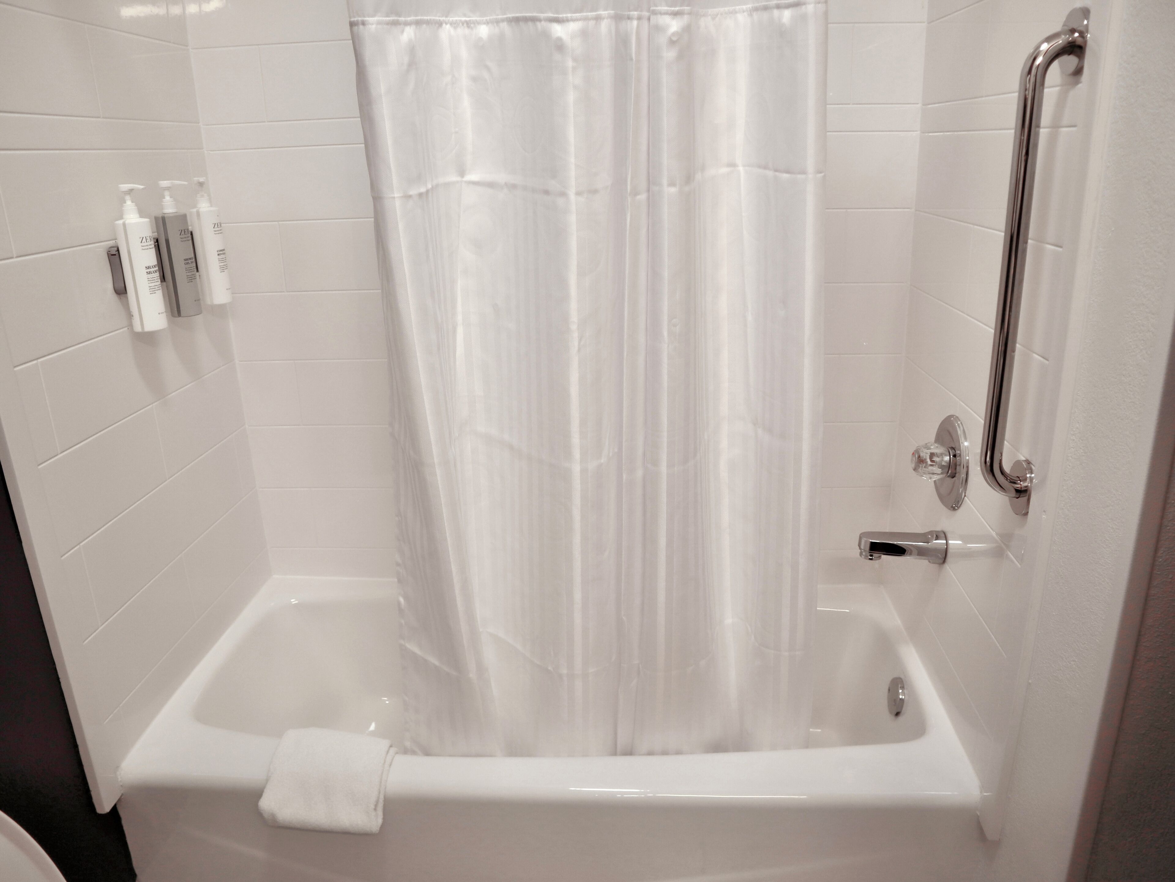 Una bañera blanca con cortina de ducha, un cabezal de ducha manual y un asidero.