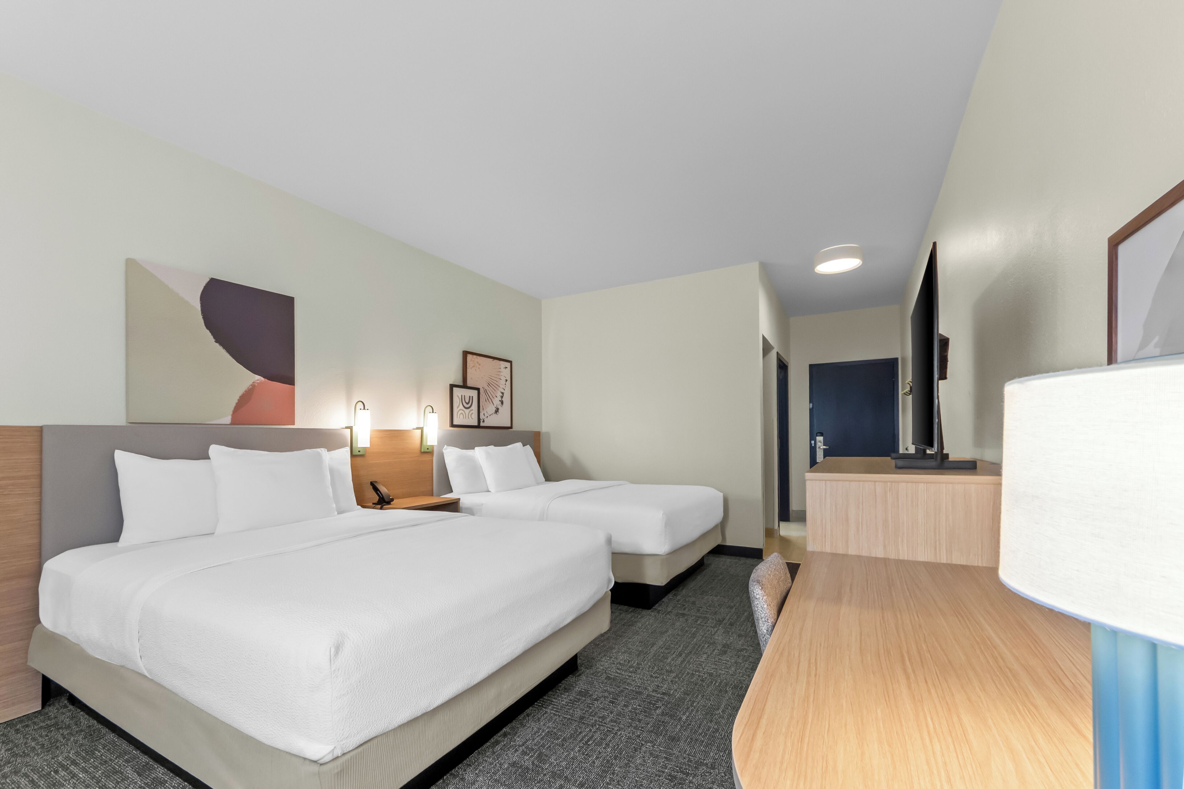A hotel room with two beds, a desk, a lamp, and a TV.