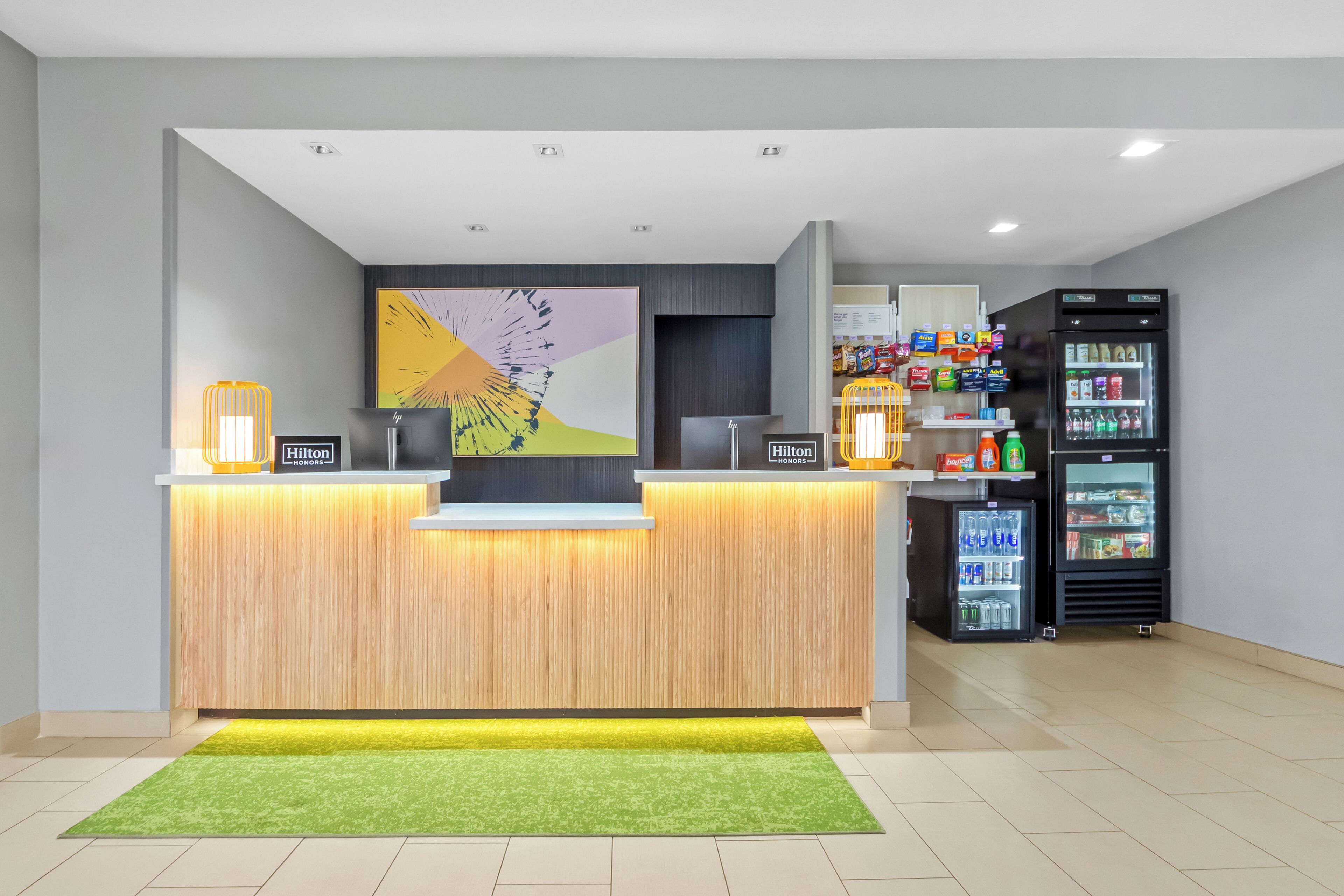 A hotel reception desk with a wooden counter, a large abstract painting, a minibar, and a refrigerator stocked with beverages.