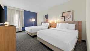 Ein Hotelzimmer mit zwei Betten, einem Schreibtisch, einem Fernseher und einem fensterbepflanzten Fenster.