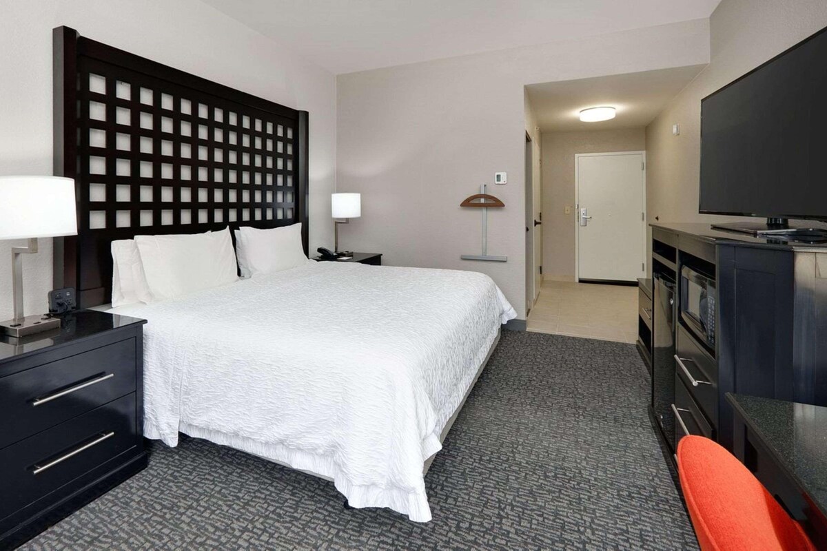 Quarto de hotel com uma cama grande, uma escrivaninha com um computador, uma televisão e um guarda-roupa.