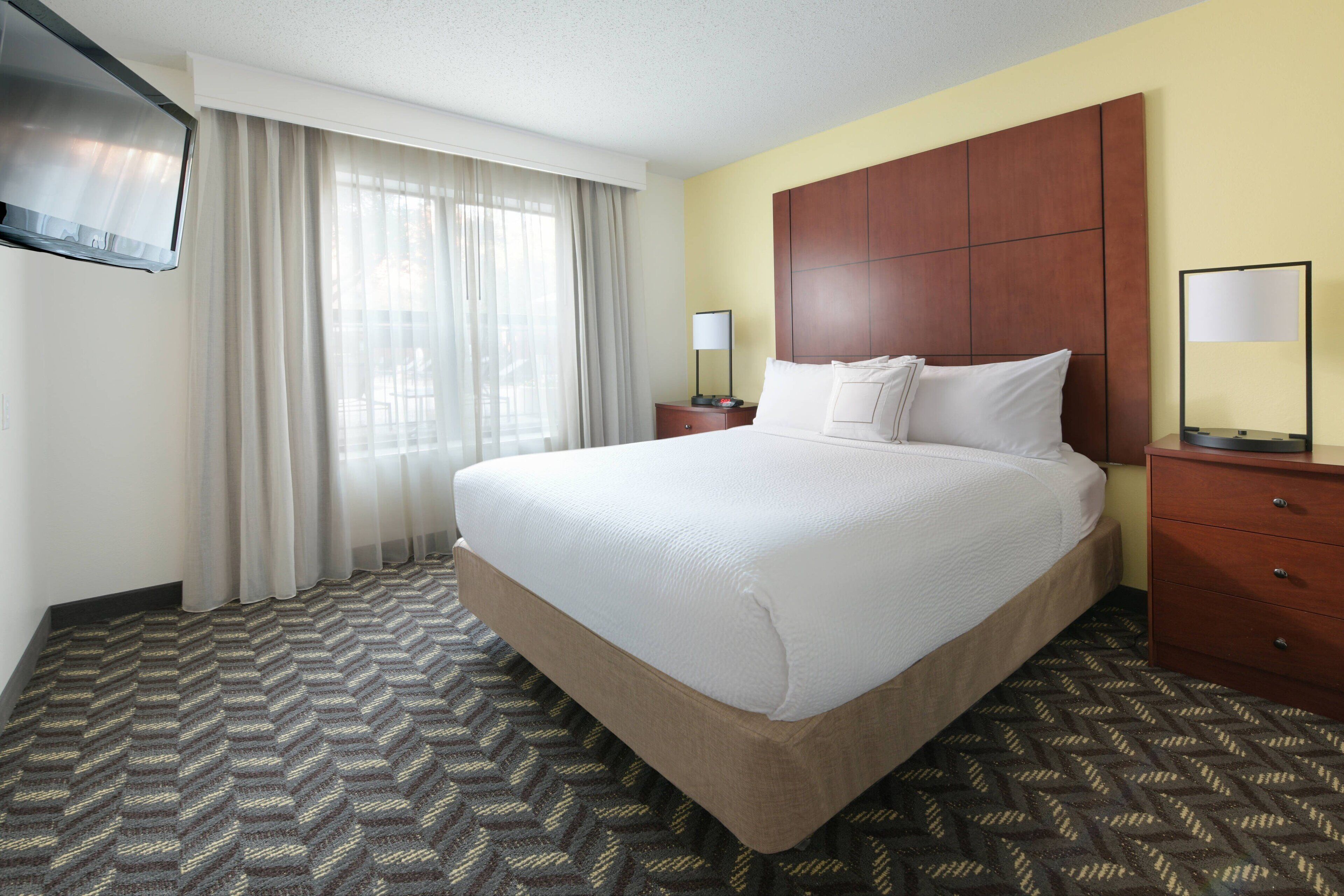 A hotel room with a large bed, a wooden headboard, two bedside tables with lamps, a flat-screen TV mounted on the wall, and a patterned carpet.