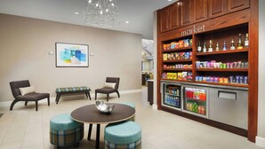 A modern lounge area with a round table, two chairs, and a bench. A refrigerator stocked with beverages and snacks is visible. - Residence Inn by Marriott Dallas Addison/Quorum Drive (Dallas)