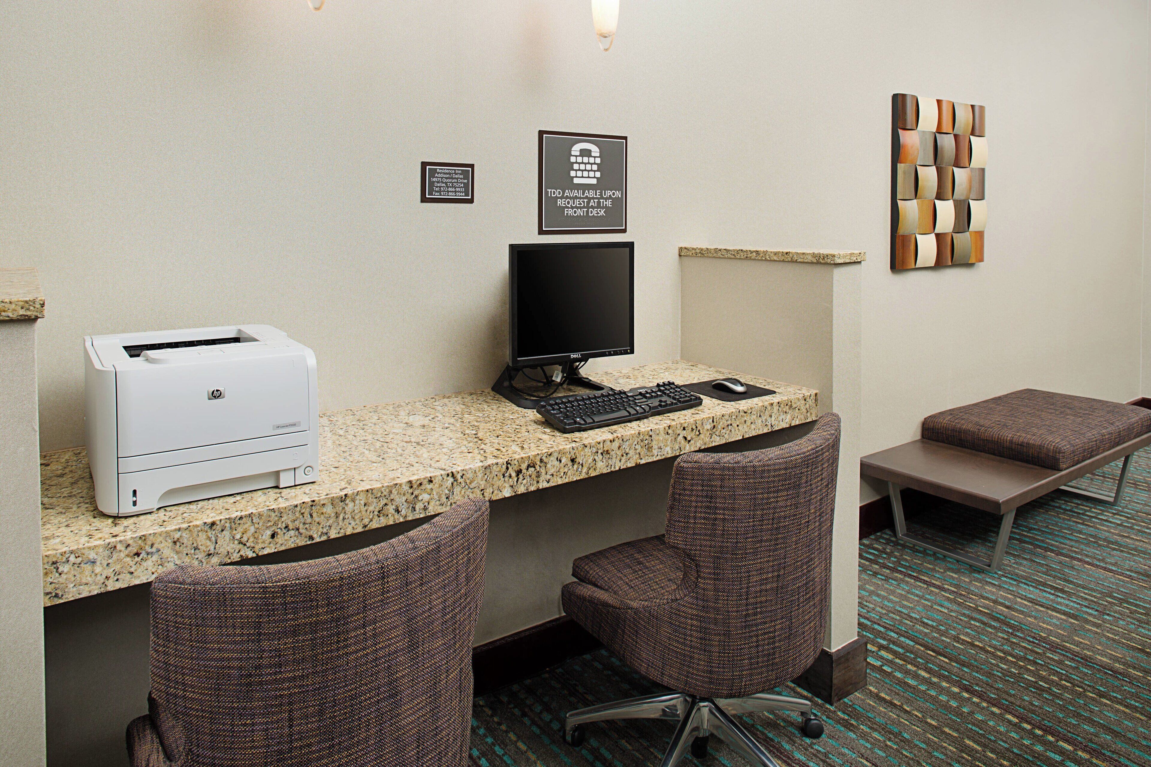 Habitación de hotel con escritorio, dos sillas, una impresora, un monitor de computadora y un teclado. Hay dos letreros en la pared y un adorno decorativo.