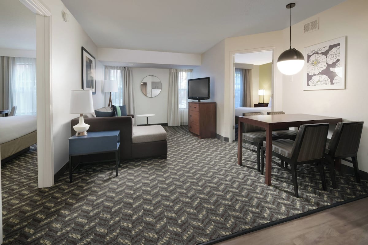 A hotel room with a sofa, a small table, a TV, a dining table with chairs, and a carpeted floor.