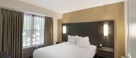 A hotel room with a large bed, a bedside table with a lamp, a window with curtains, and a view of trees outside.