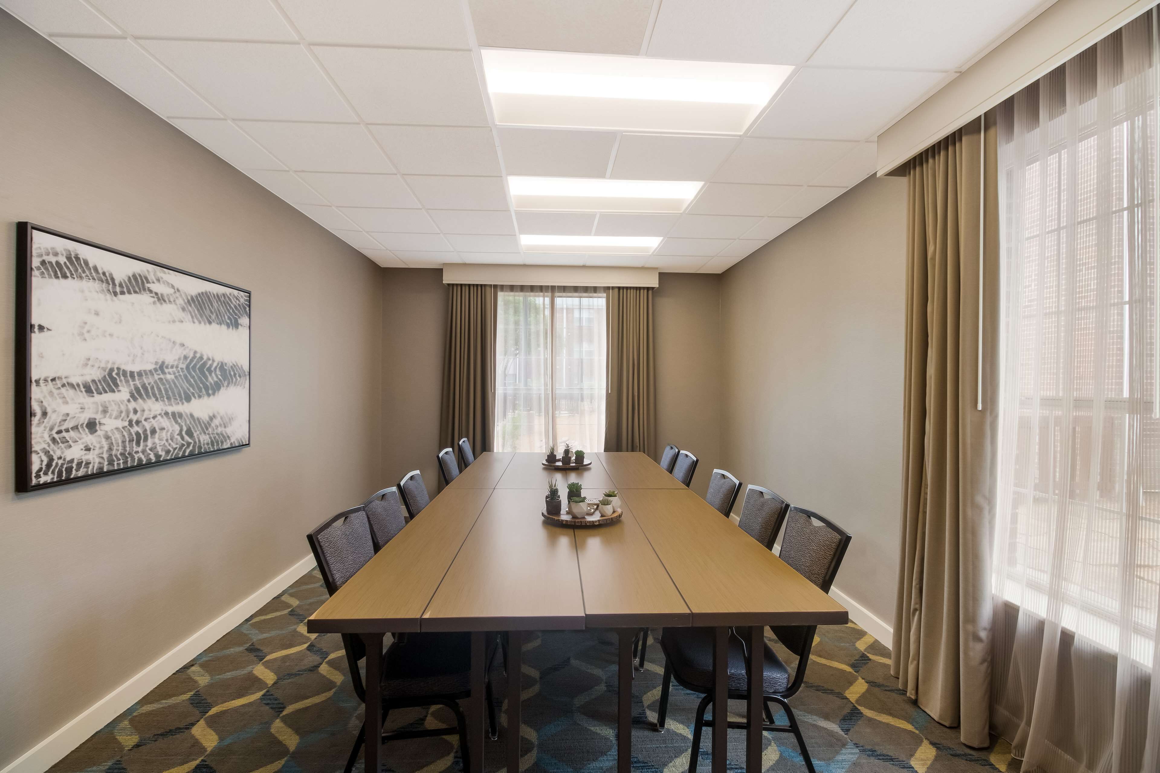 A conference room with a long table, chairs, a framed picture on the wall, and a window with curtains.