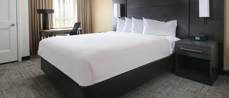 A hotel room with a large bed, a desk, a chair, a nightstand with a clock, and a window with curtains.