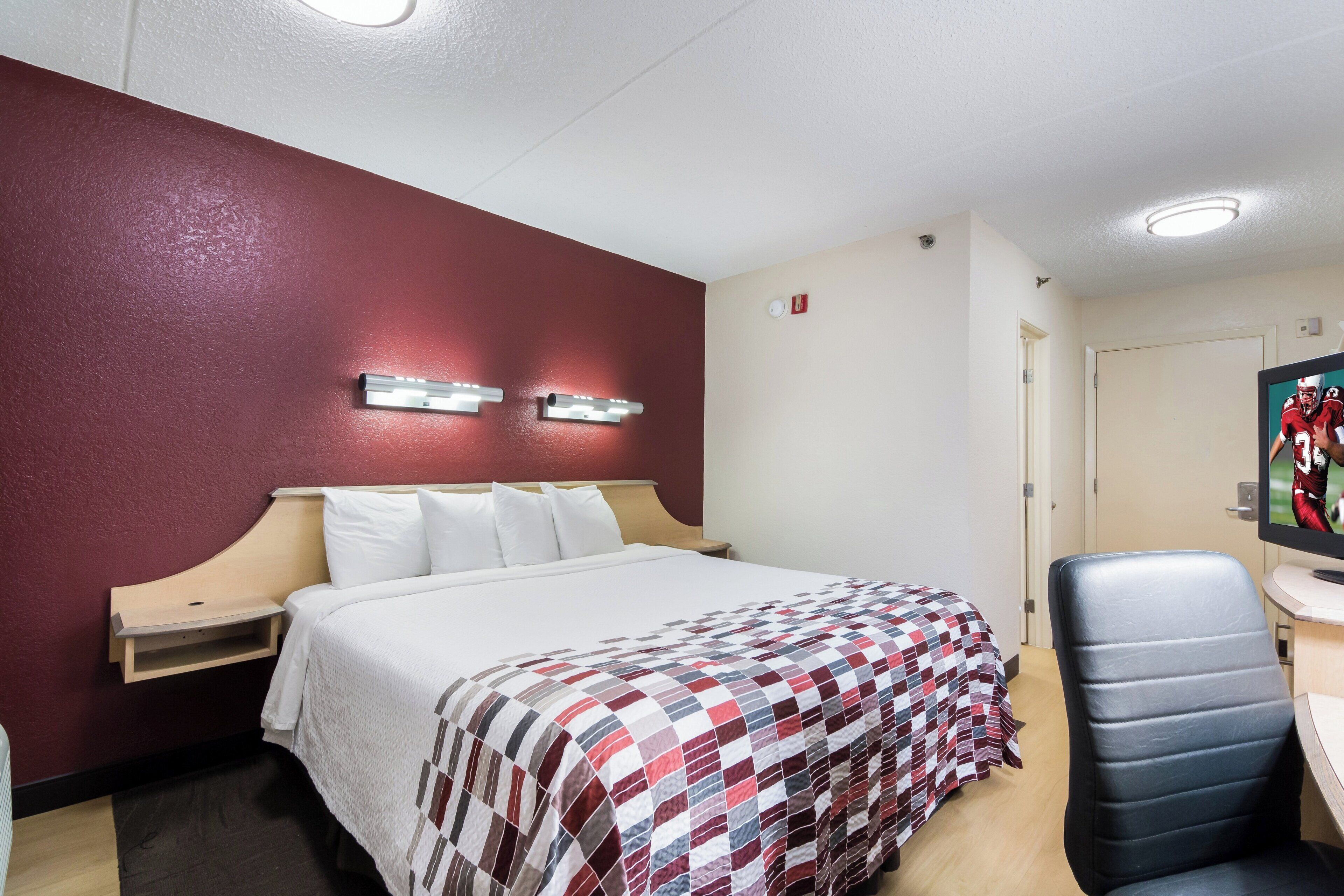 A hotel room with a bed, a desk with a chair, a wall-mounted light fixture, and a door.