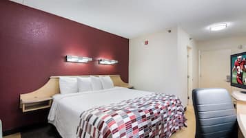 A hotel room with a bed, a desk with a chair, a wall-mounted light fixture, and a door.